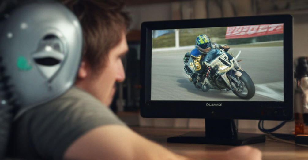 A man sits with a beer, eyes fixed on his computer monitor showing a motorcycle race.