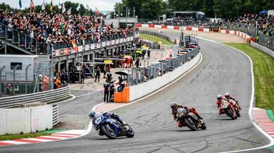 Professional motorcycle racers competing on a winding circuit, dramatic track environment, pit crew and spectators in the background